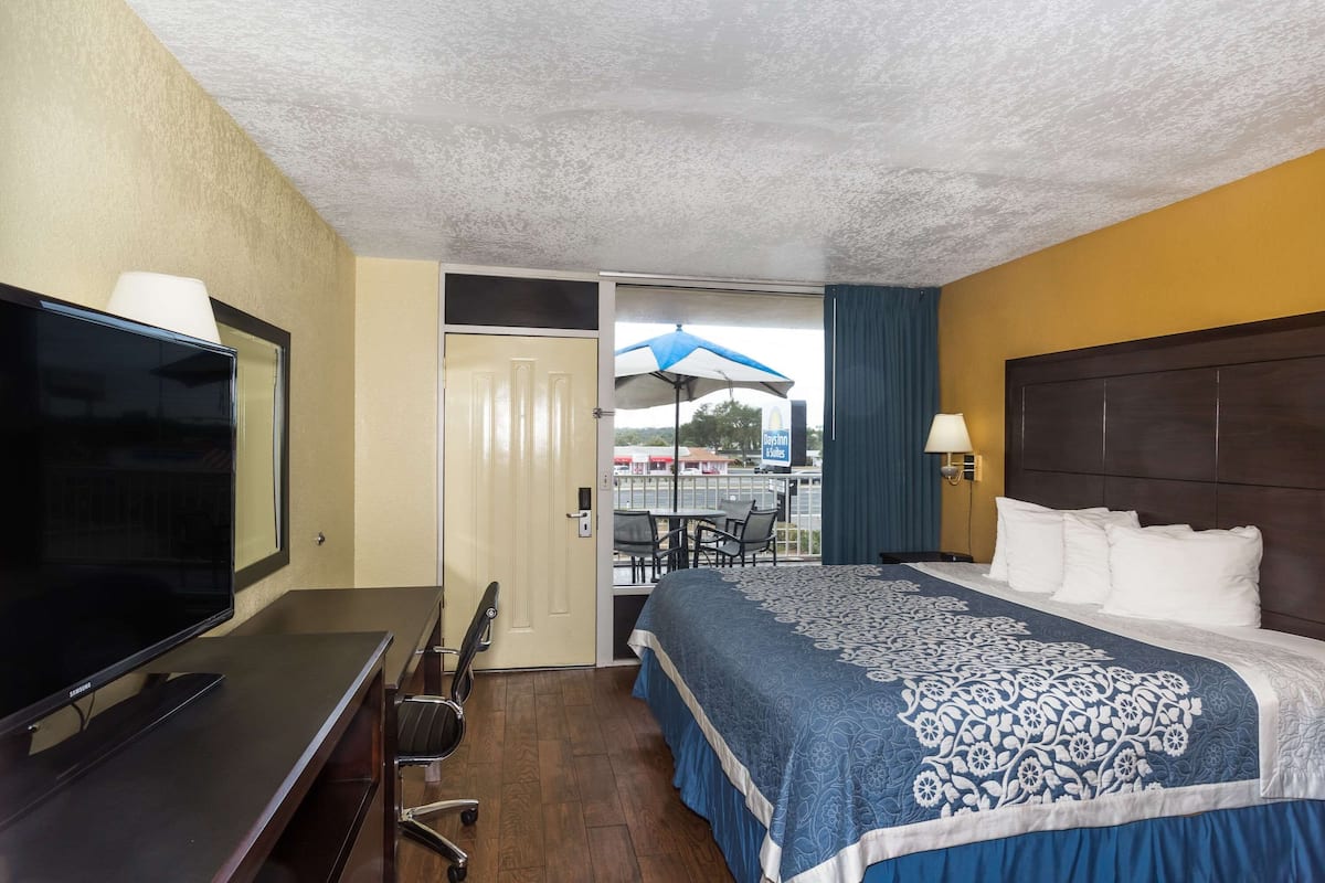 A hotel room with a bed, a desk with a TV, a chair, and a view of an outdoor area with a table and umbrella.