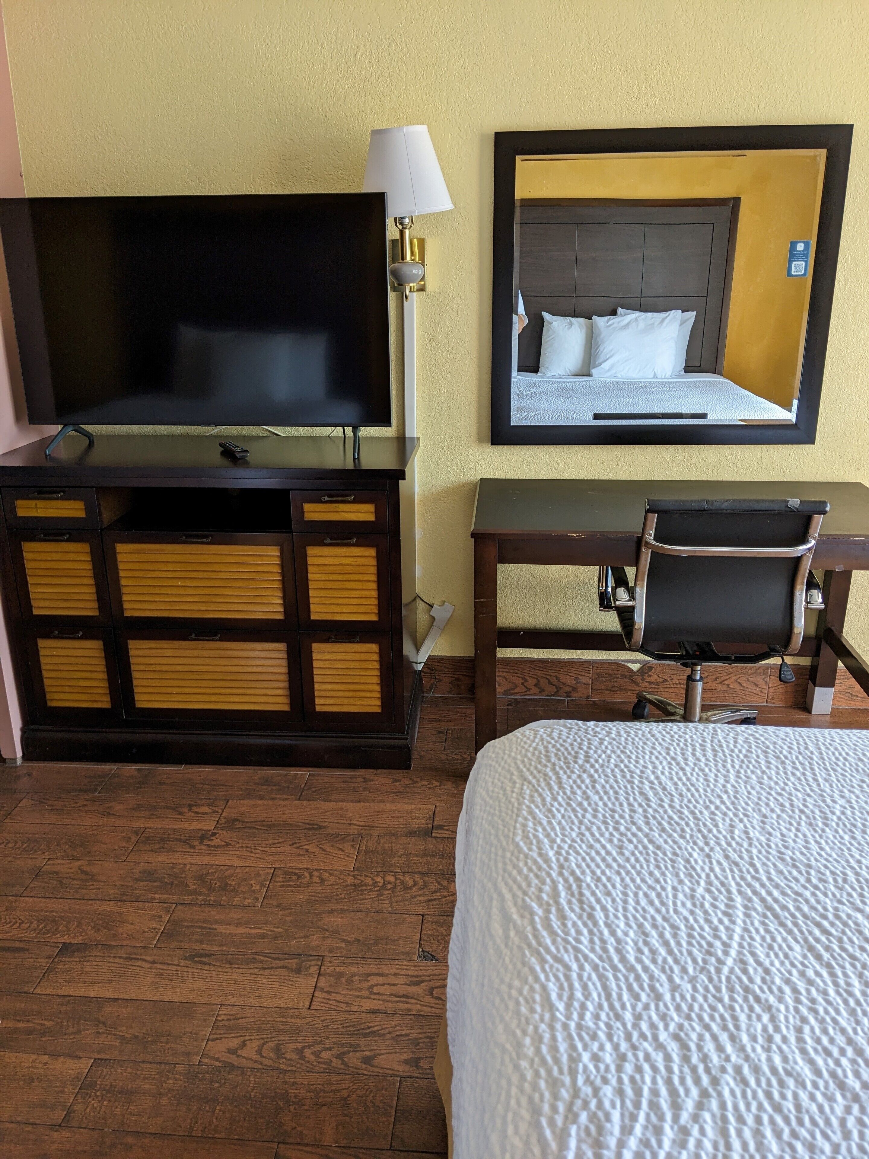 Habitación de hotel con cama, escritorio con silla, televisión y un espejo que refleja una cama.