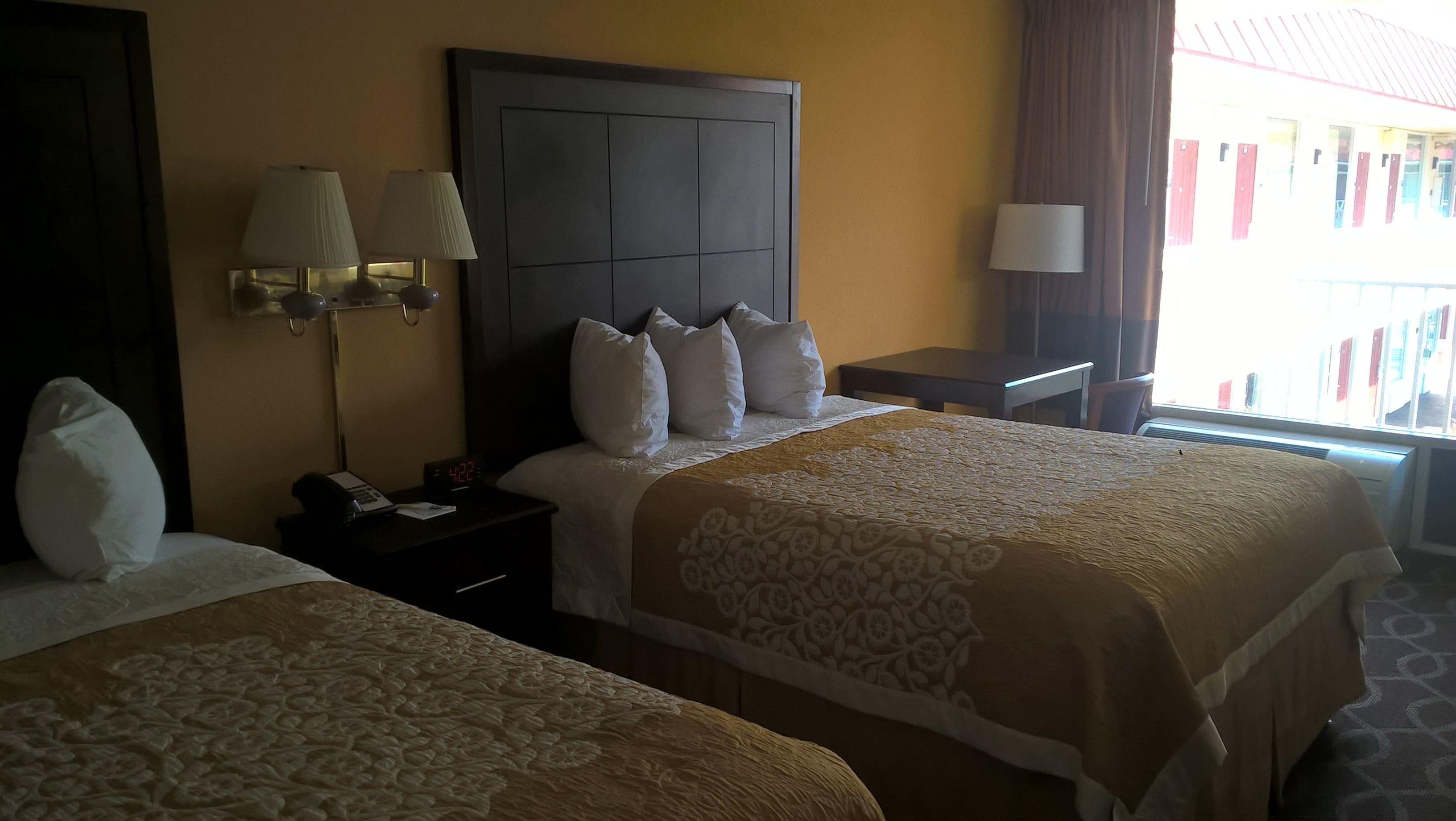 A hotel room with two beds, a nightstand with a lamp, a clock, and a window with curtains.