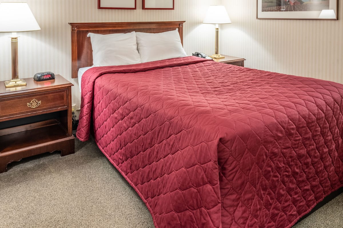 Une chambre d’hôtel avec un grand lit, deux tables de chevet, une petite table d’appoint, une couvre-lit rouge matelassée, deux tableaux encadrés au mur et une lampe sur chaque table de chevet.