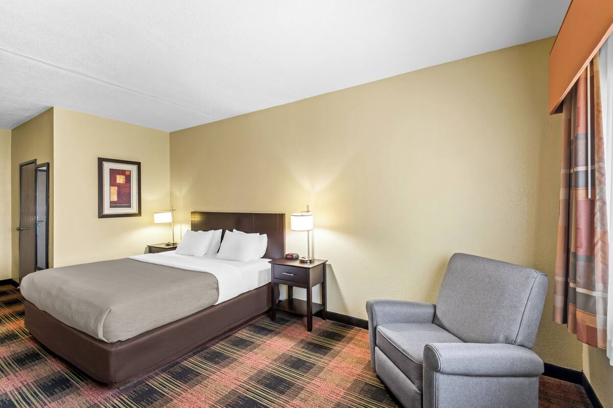 A hotel room with a large bed, a chair, a nightstand with a lamp, and a framed picture on the wall.