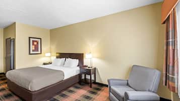 Quarto de hotel com uma cama grande, uma cadeira, uma mesinha de cabeceira com abajur e um quadro na parede.