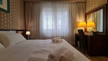 Habitación de hotel con una cama bien hecha, dos toallas enrolladas, un escritorio con una silla, una lámpara y un espejo grande.