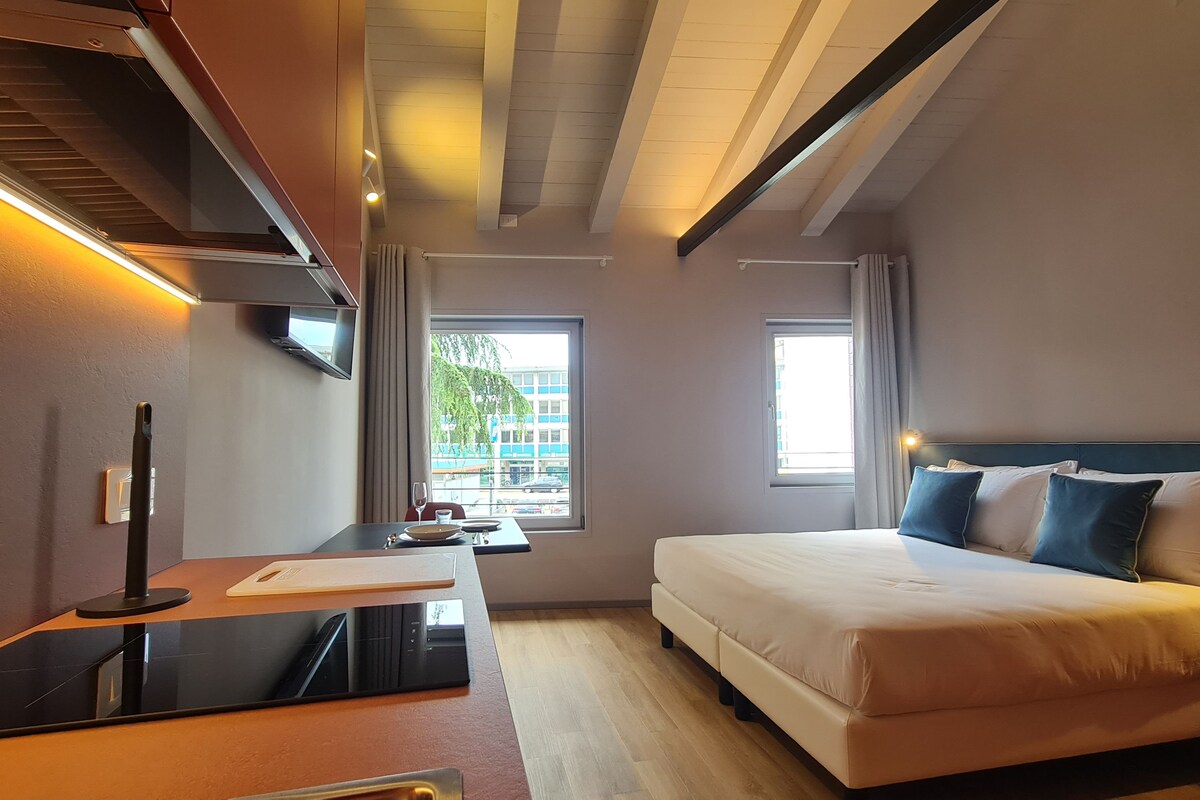 A hotel room with a bed, a desk with a computer, a TV, and a window with a view of buildings.