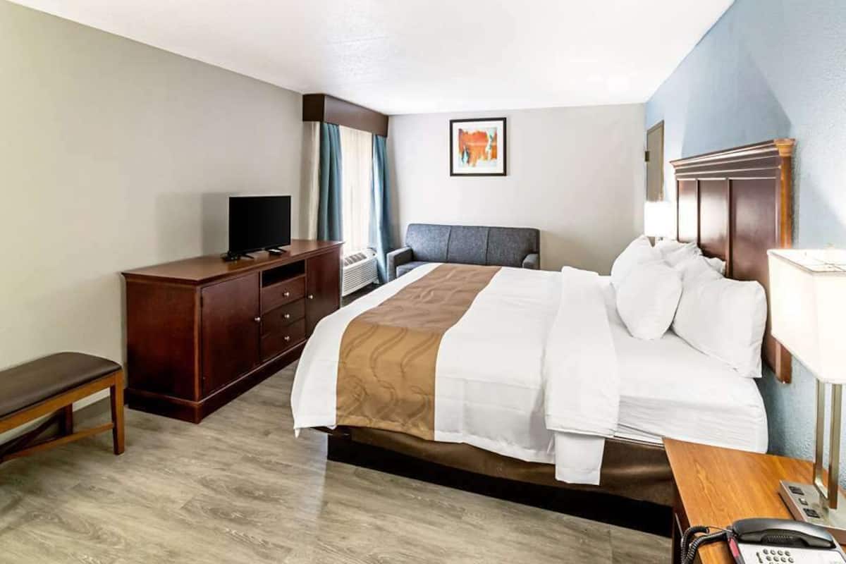 A hotel room with a bed, a wooden dresser, a bench, a desk with a phone, and a TV.