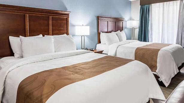 A hotel room with two beds, wooden headboards, white bedding, a nightstand with a lamp, and a window with curtains. - Farragut Inn & Suites (Farragut)