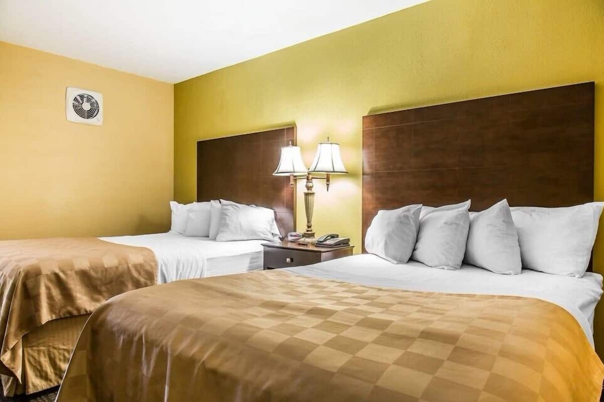 Habitación de hotel con dos camas, cabecero de madera, mesita de noche con lámpara y ventilador de pared.