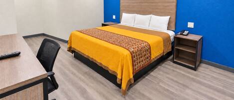 A hotel room with a bed, bedside table, chair, and a desk with a remote control.