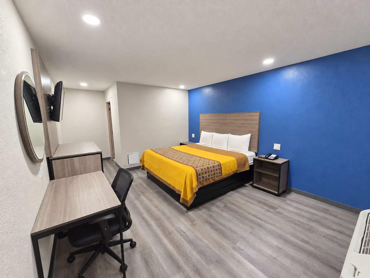 A hotel room with a bed, desk, chair, bedside table, and a mirror.