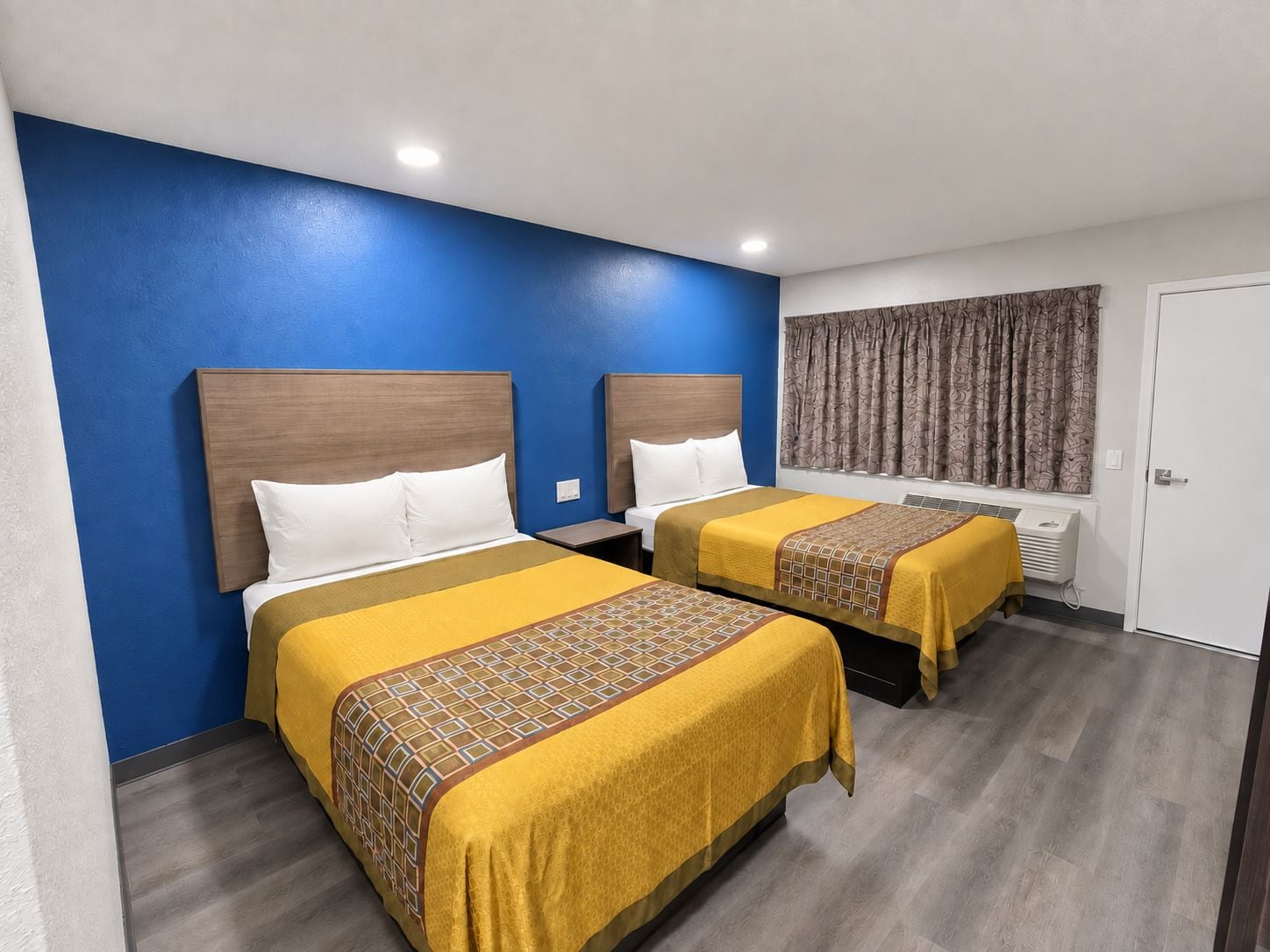 A hotel room with two beds, each with a wooden headboard, white pillows, and yellow bedspreads with a geometric pattern.
