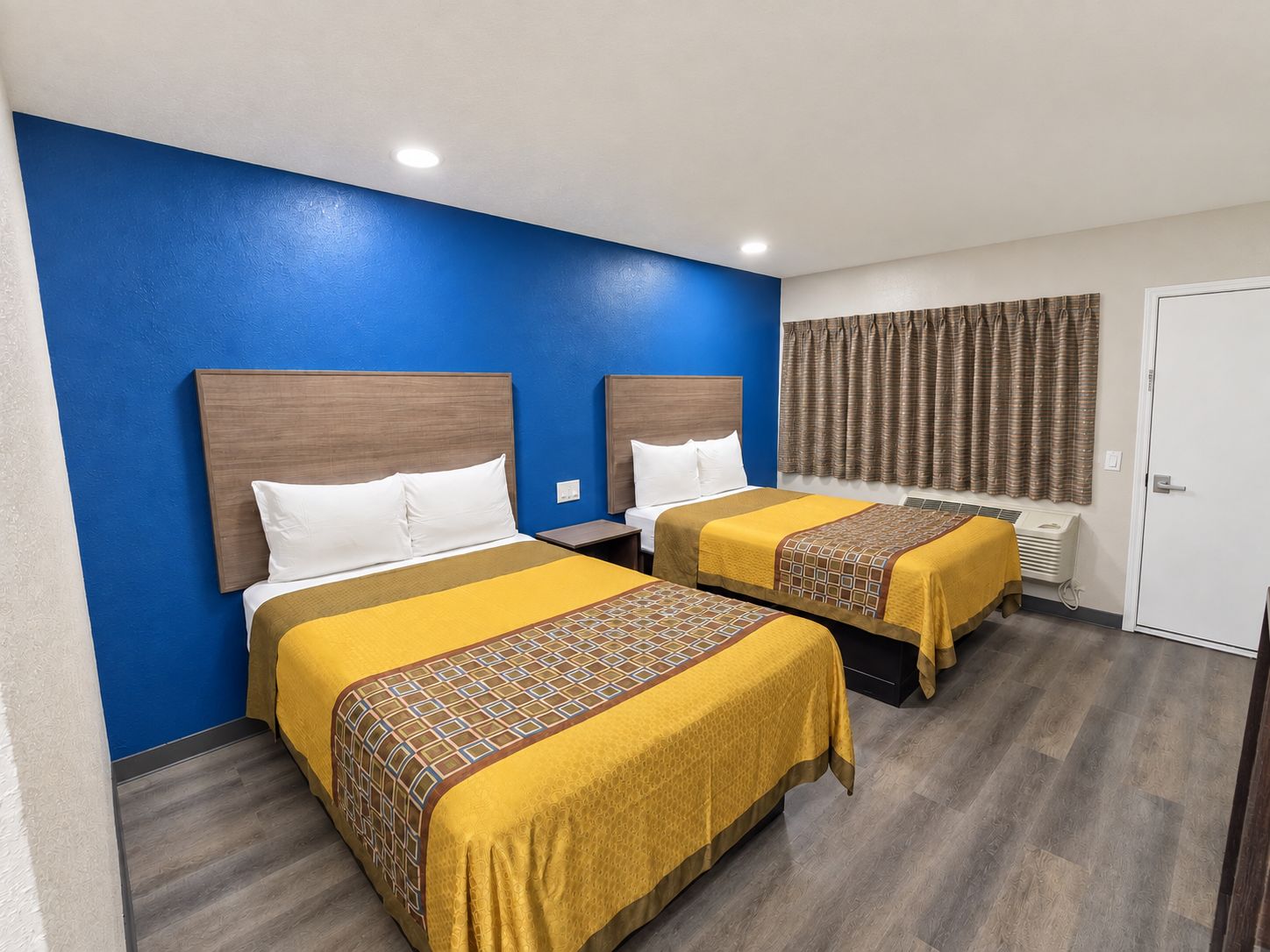 A hotel room with two beds, a blue accent wall, and a window with curtains.