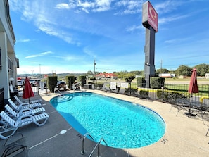 A swimming pool with lounge chairs and umbrellas, surrounded by a fence, with a view of a hotel and distant buildings.