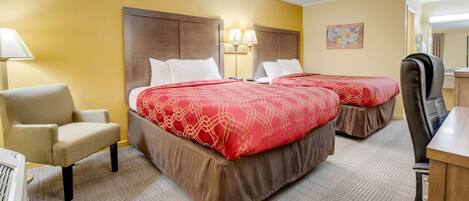 A hotel room with two beds, a desk, a chair, and a lamp.