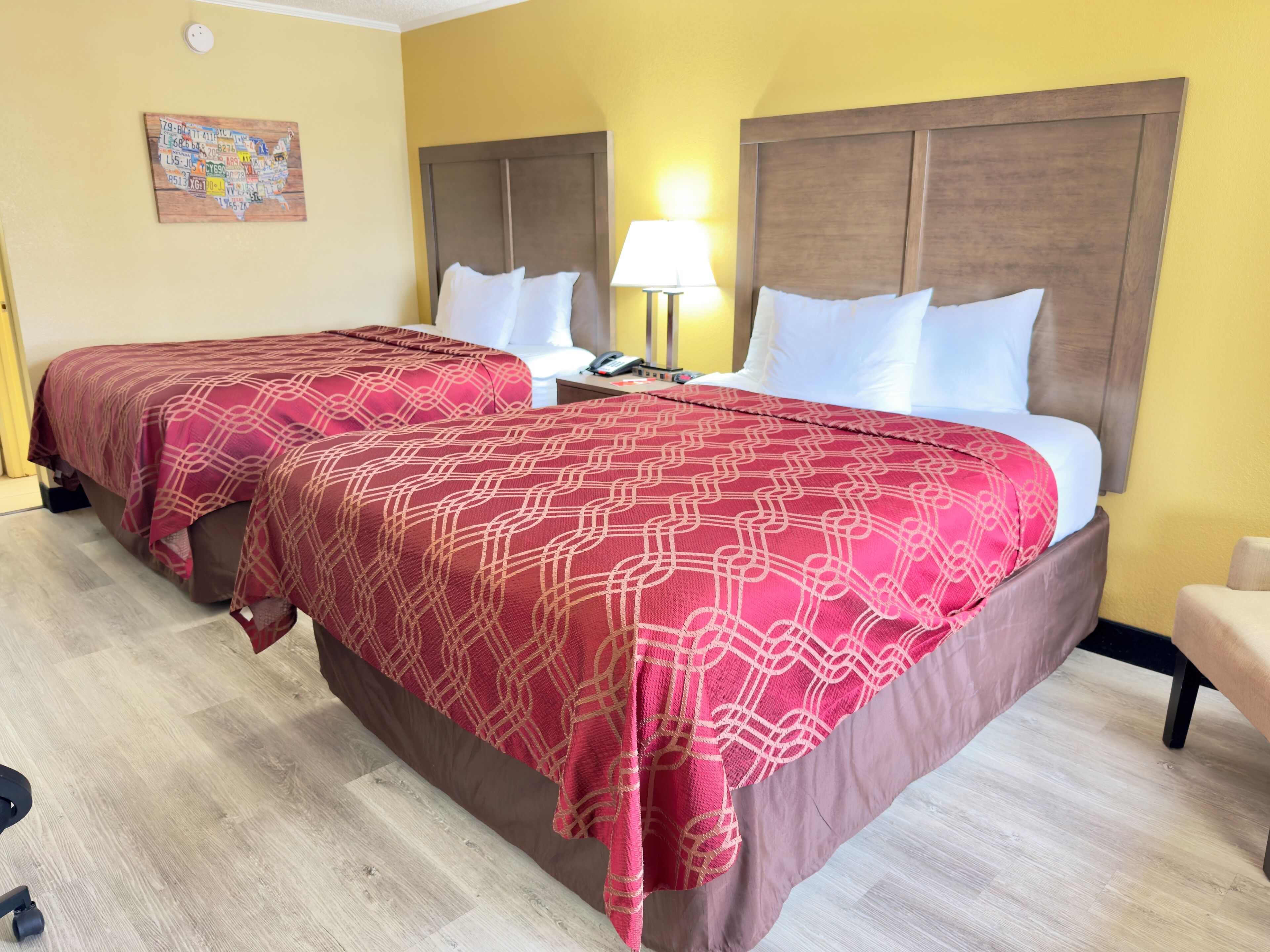Une chambre d’hôtel avec deux lits, chacun recouvert d’une couvre-lit à motifs rouges et agrémenté d’oreillers blancs. On y trouve une table de chevet avec une lampe et un téléphone, ainsi qu’une horloge murale.