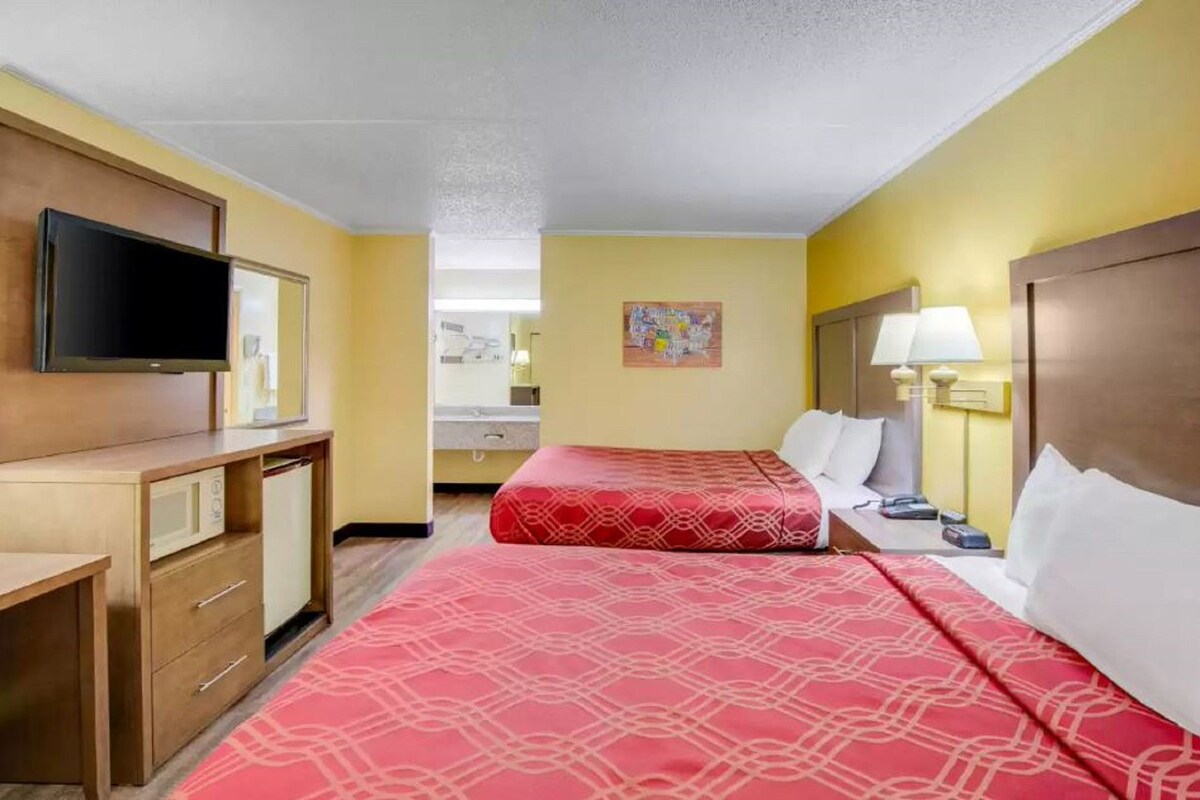 A hotel room with two beds, a TV, a desk, and a bathroom visible in the background.