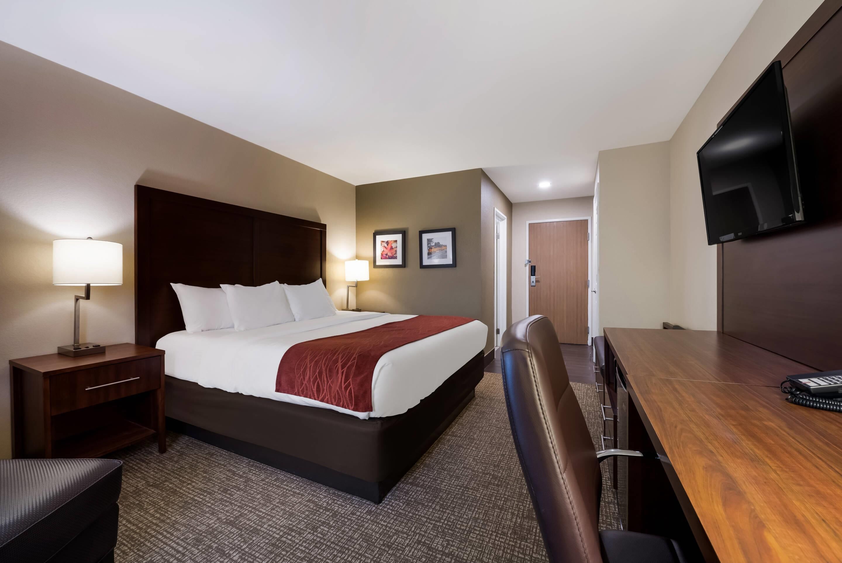 Una habitación de hotel con cama, mesita de noche con lámpara, escritorio con silla, televisor montado en la pared y dos cuadros enmarcados.