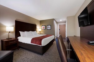 Una habitación de hotel con cama, mesita de noche con lámpara, escritorio con silla, televisor montado en la pared y dos cuadros enmarcados.
