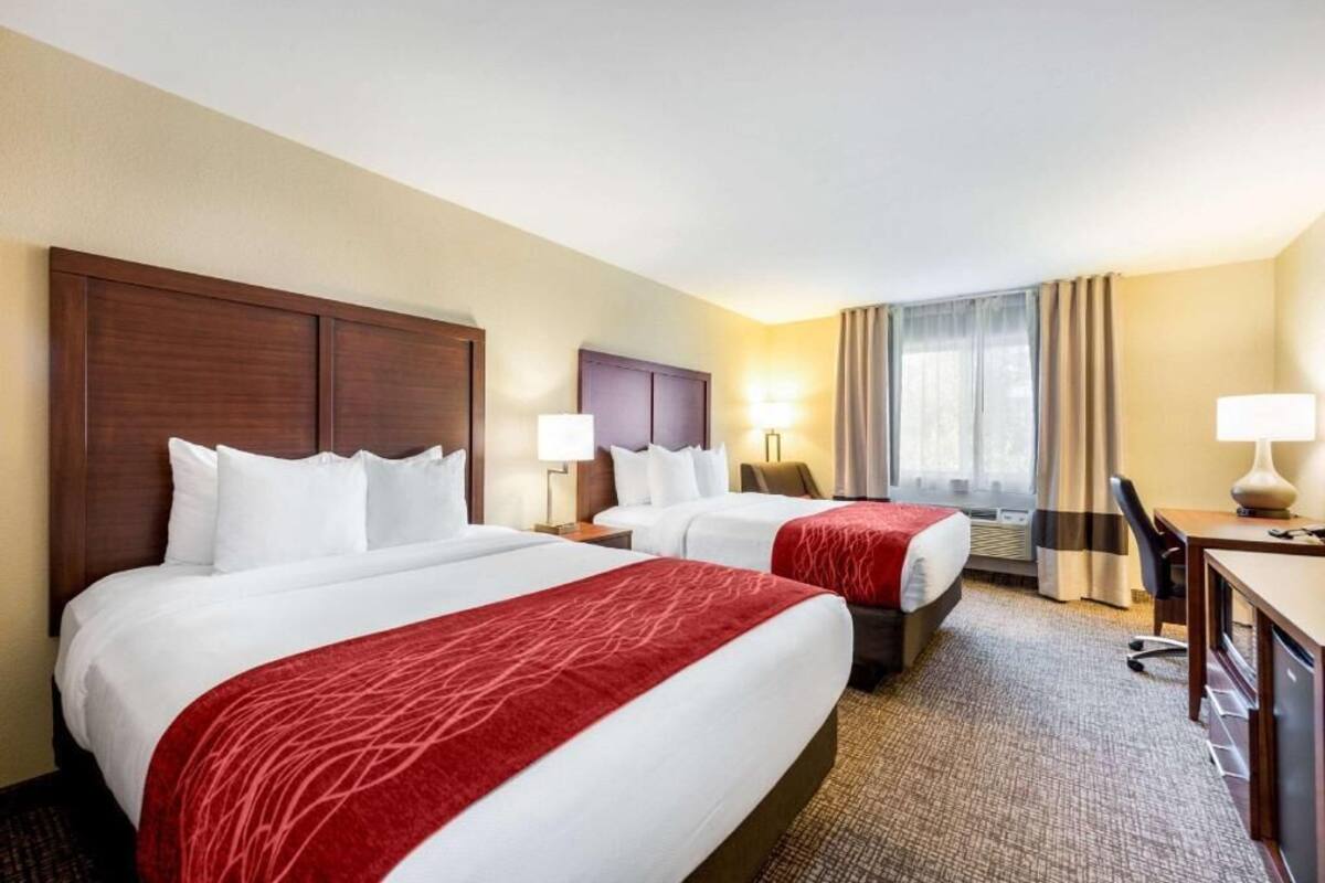 A hotel room with two beds, a desk, a chair, and a lamp.