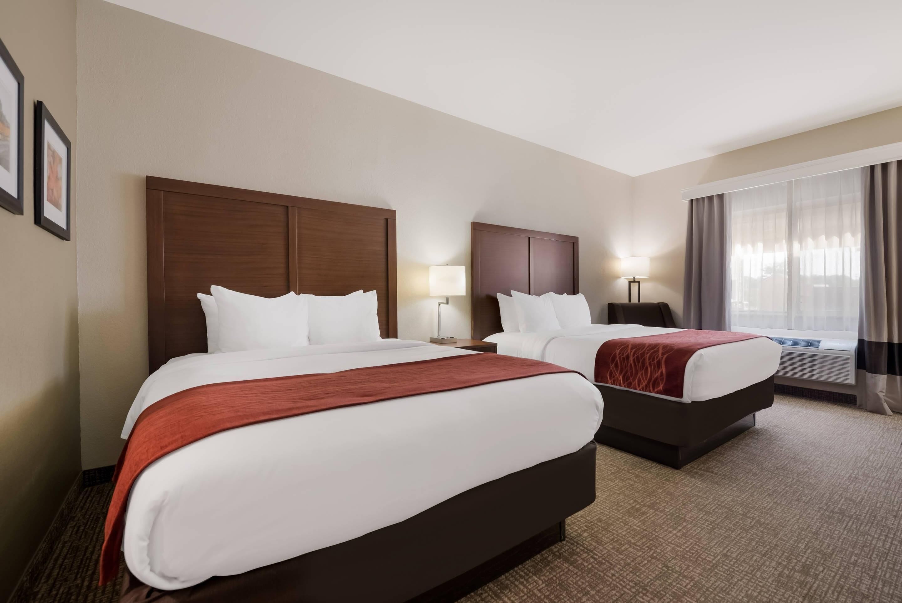 Habitación de hotel con dos camas, cada una con cabecero de madera, ropa de cama blanca y una manta roja. Hay una mesita de noche con lámpara entre las camas, y un ventanal con cortinas a la derecha.