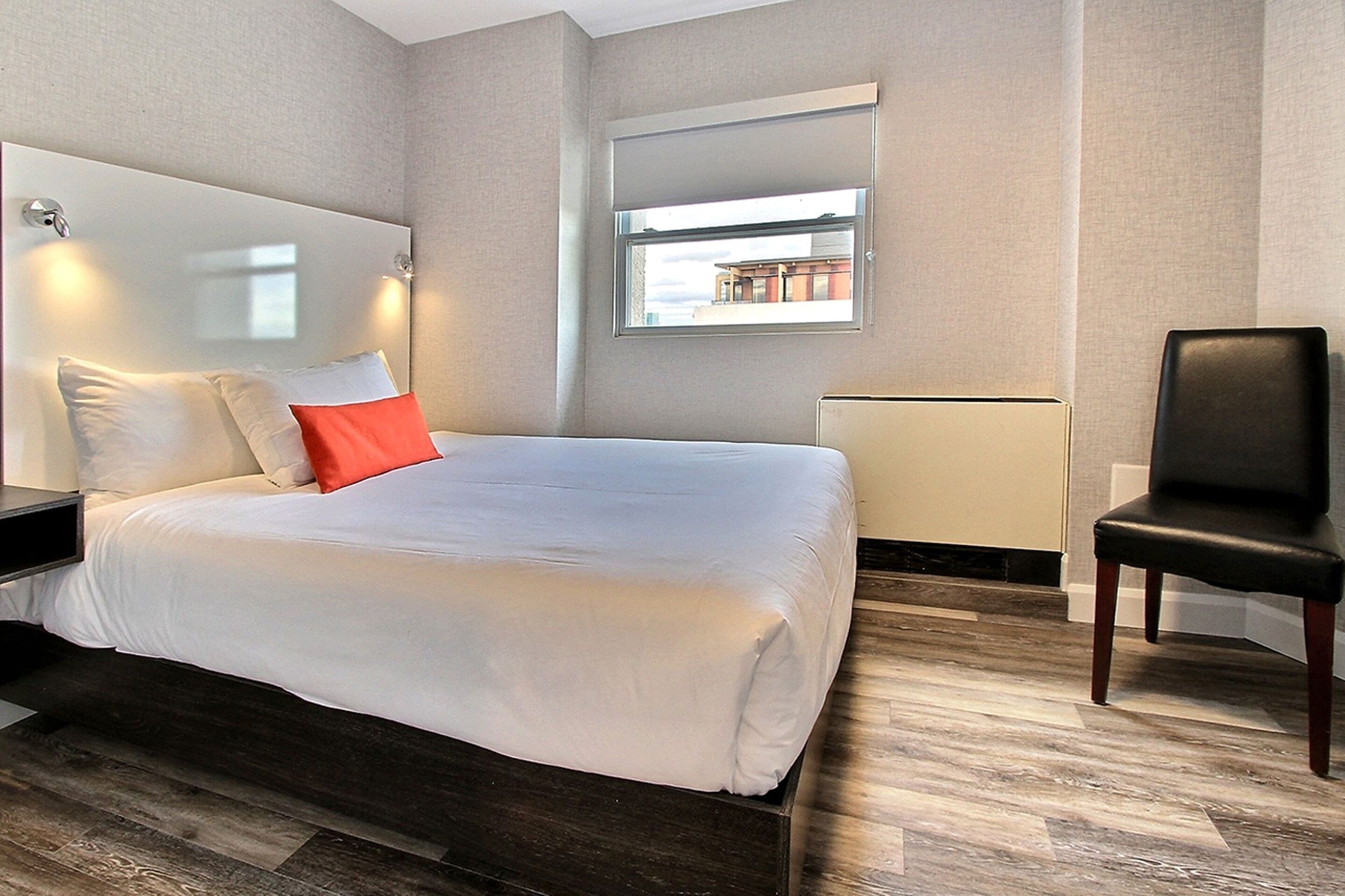 A modern hotel room with a large bed, a chair, a window with a view, and a mirror on the wall.