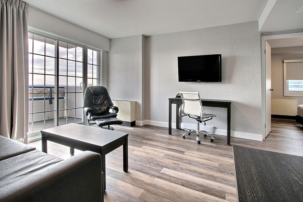 A modern hotel room with a large window, a flat-screen TV mounted on the wall, a leather chair, a desk with a chair, and a sofa.