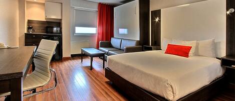 Una habitación de hotel moderna con una cama grande, un escritorio con silla, una kitchenette pequeña y un televisor.
