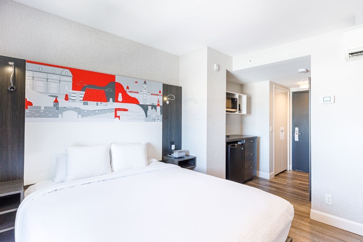 Habitación de hotel con una cama grande, un mural de la ciudad, una kitchenette pequeña y un baño separado.