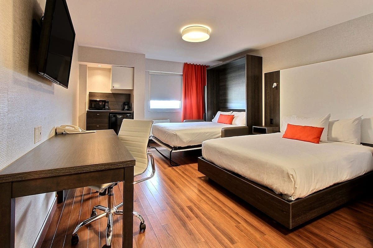 A hotel room with a wooden floor, two beds, a desk, a chair, a TV, a kitchen area, and a window with red curtains.