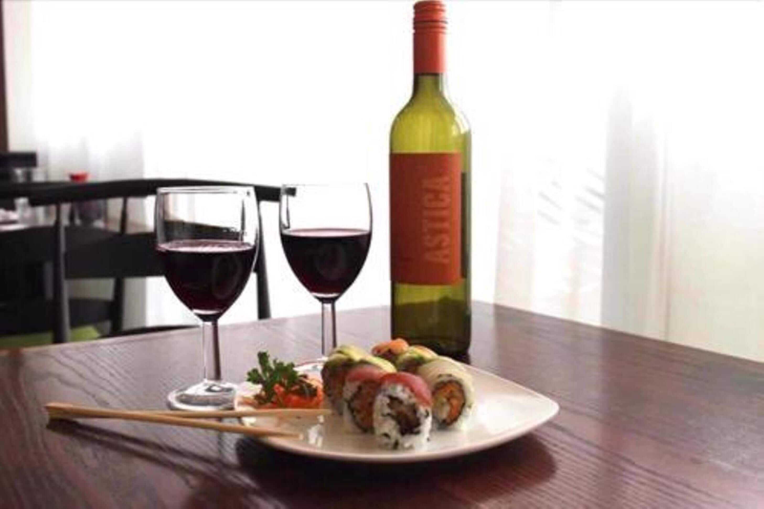 A table with sushi, two glasses of red wine, and a bottle of wine.