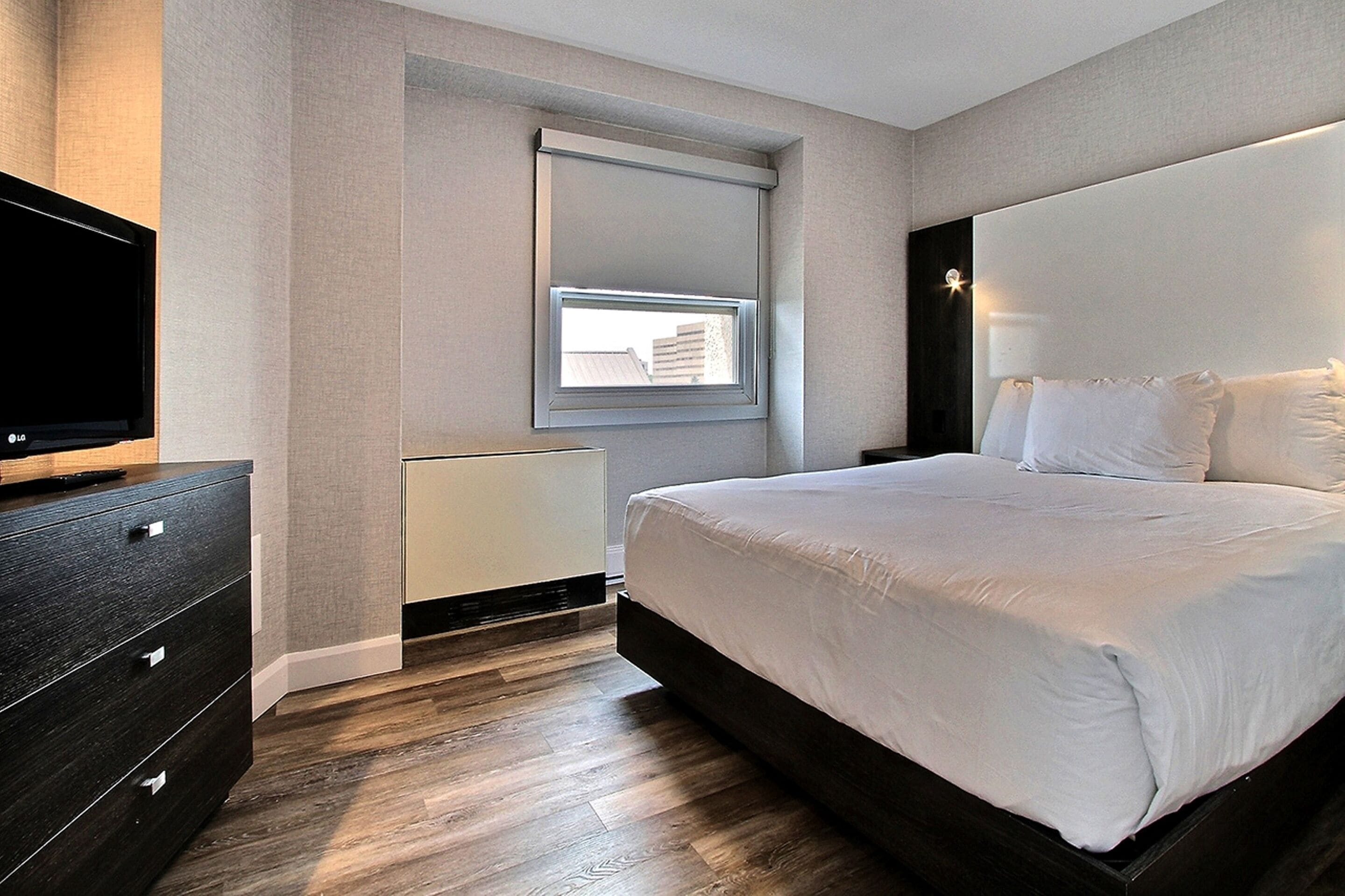 A modern bedroom with a large bed, a TV, a window with blinds, and a radiator.