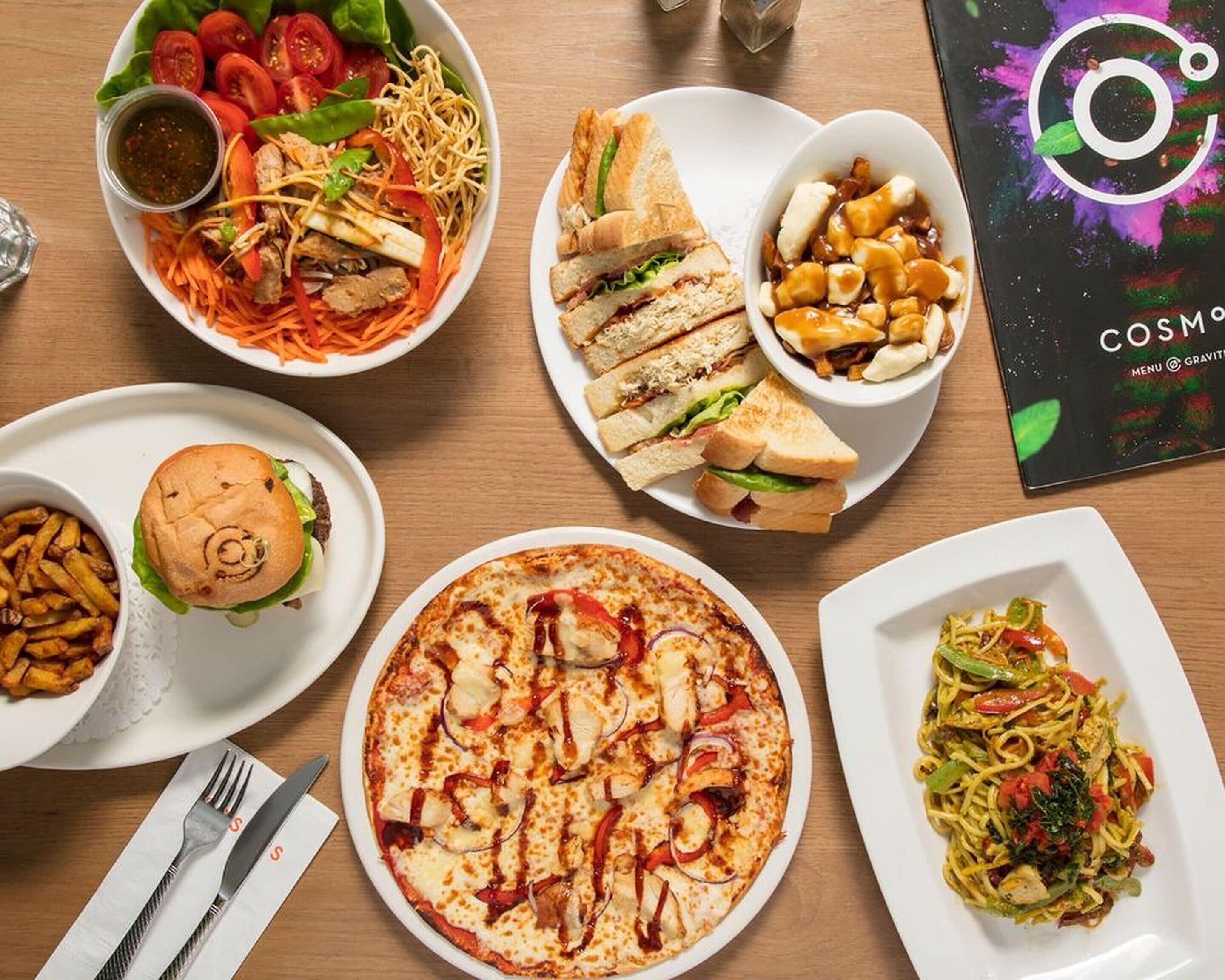 A variety of dishes including a burger, pizza, and pasta, arranged on a wooden table.