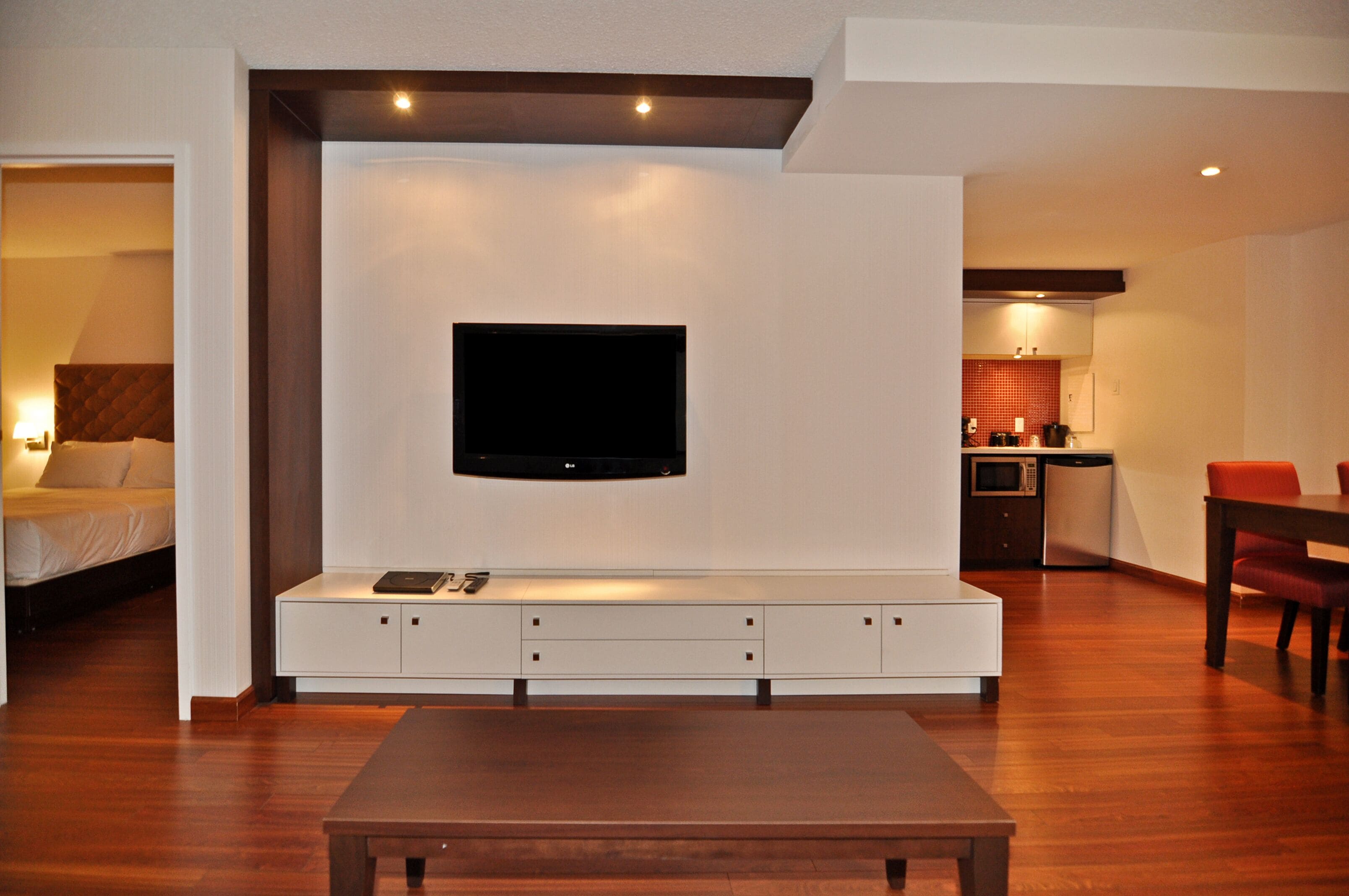 A modern living room with a flat-screen TV mounted on a white wall, a wooden coffee table, and a dining area with a red chair.