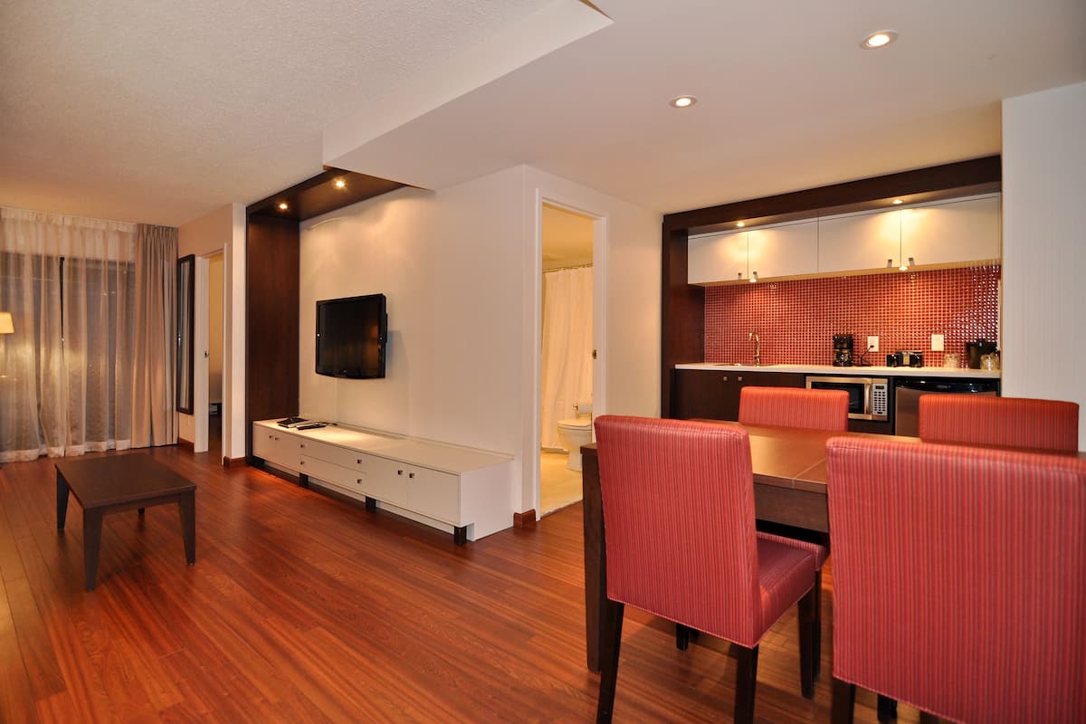 A modern hotel room with a wooden floor, a flat-screen TV mounted on the wall, a dining area with red upholstered chairs, and a kitchenette with red tiled backsplash.