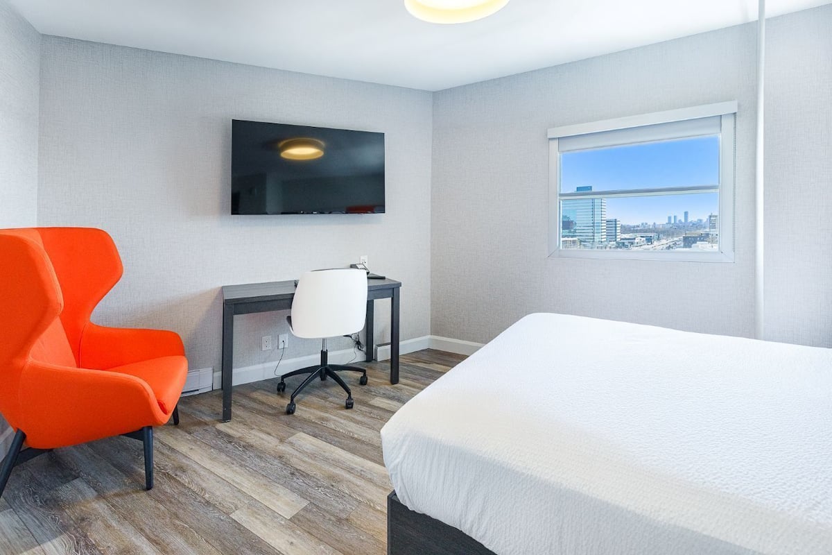 A hotel room with a bed, a desk with a chair, a TV mounted on the wall, and a window with a city view.