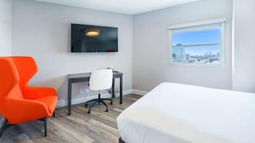 A hotel room with a bed, a desk with a chair, a TV mounted on the wall, and a window with a city view.