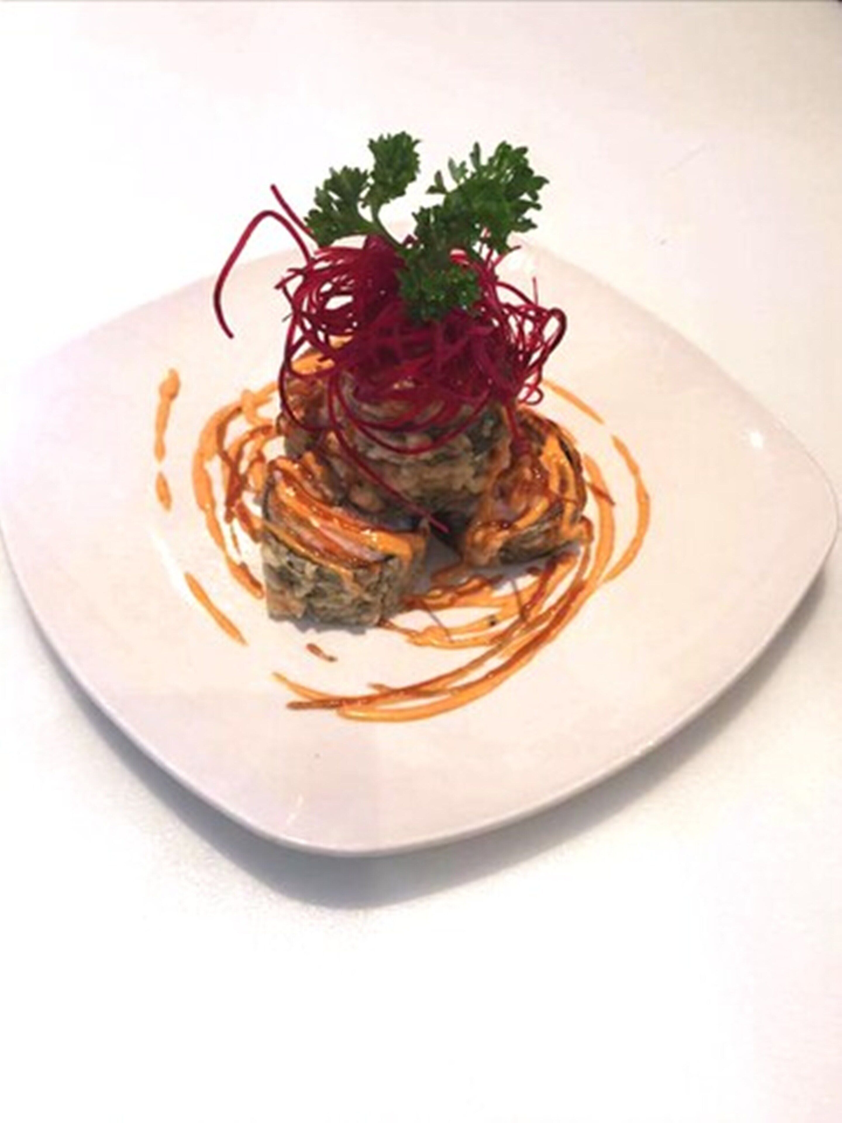 Un plato cuadrado con un guiso de ingredientes en capas, adornado con vegetales rojos y hierbas verdes.