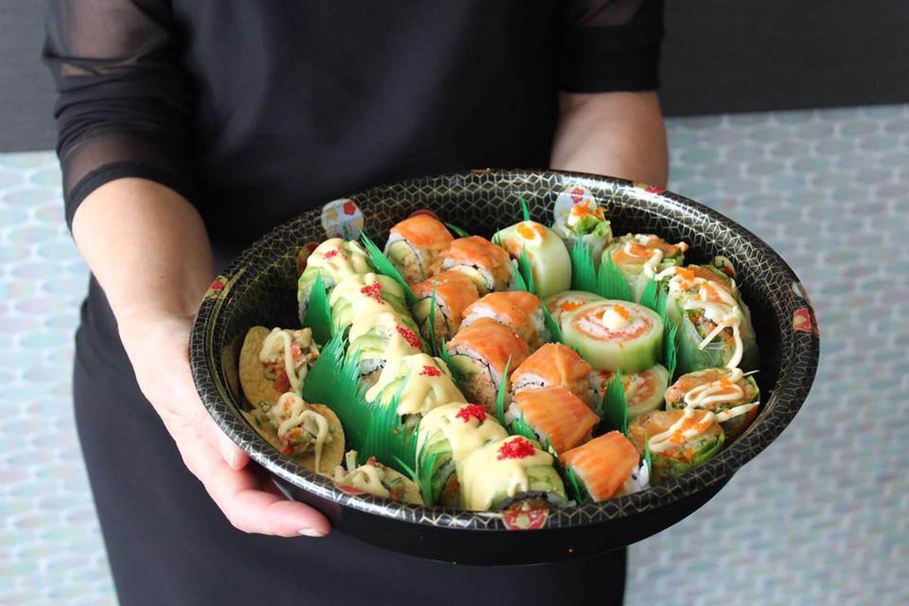 Eine Person hält eine Tablett mit verschiedenen Sushi-Rollen und Vorspeisen.