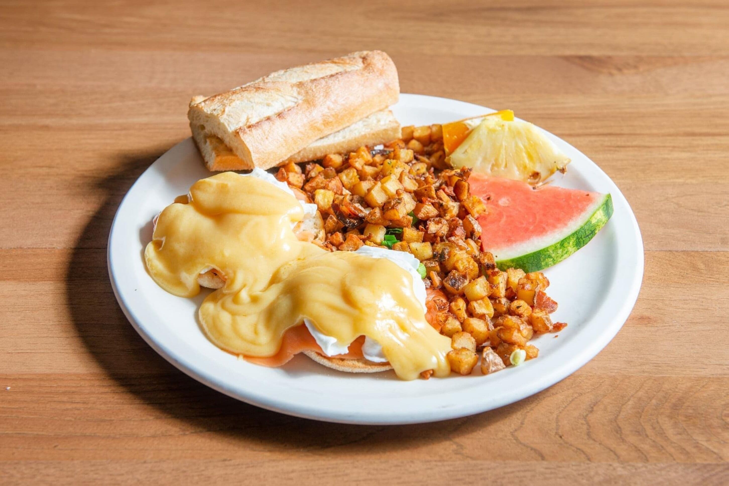 Un plato con huevos benedictinos, patatas ralladas, una rebanada de sandía y una tostada.