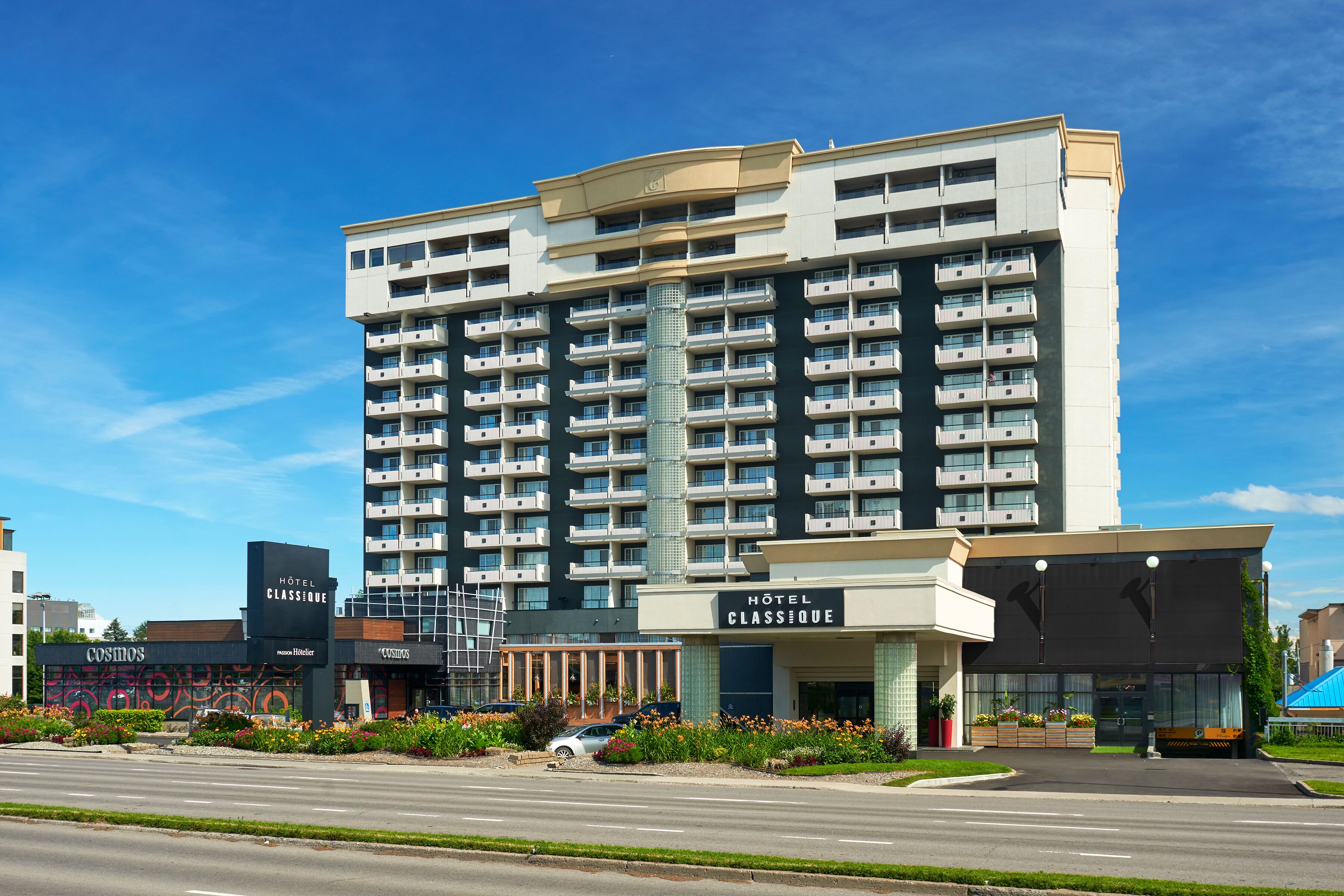 A multi-story hotel with a modern design, featuring balconies on each floor, a prominent entrance, and a landscaped area with flowers.