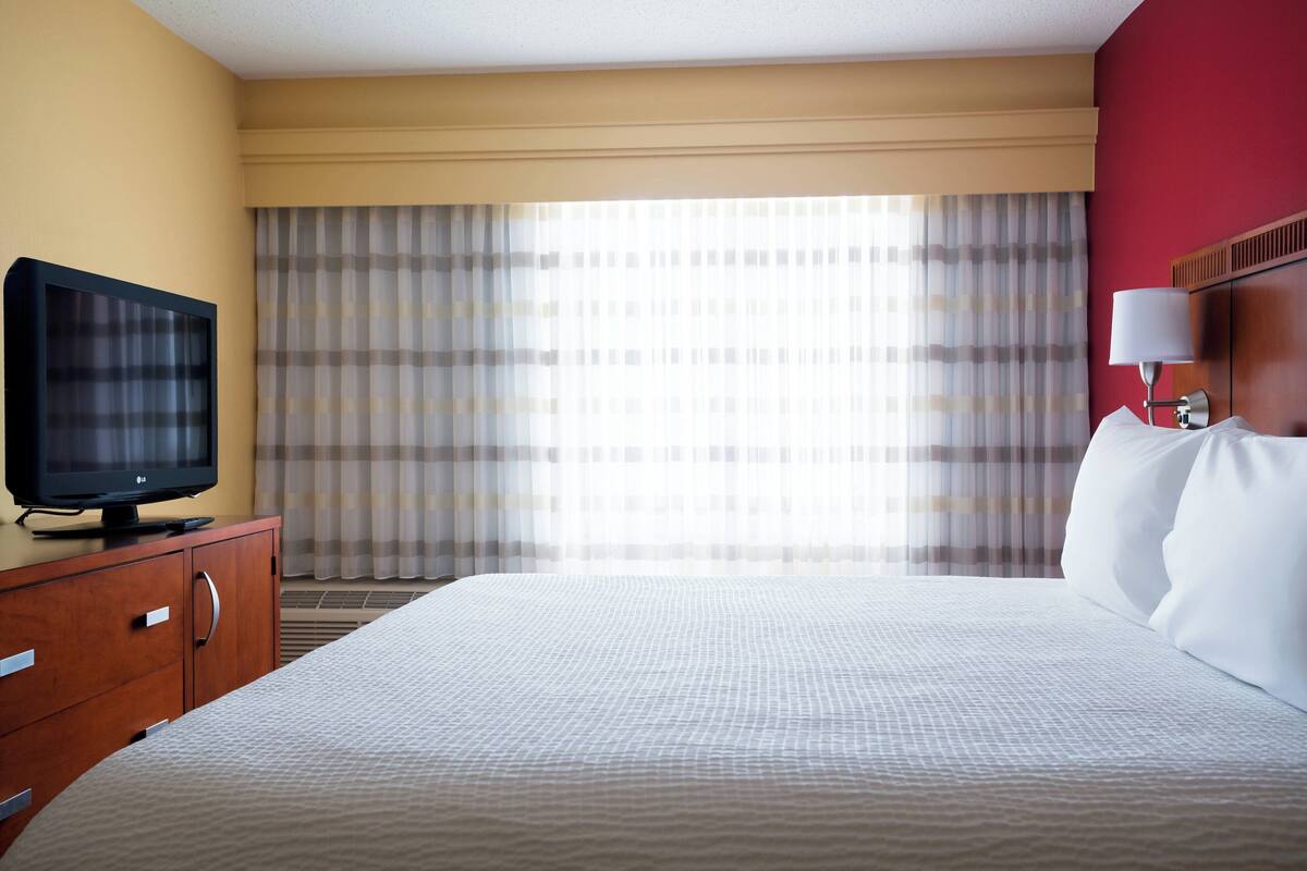 Una camera d'albergo con un letto, una televisione su una cassettiera in legno, una lampada e una finestra con tende.
