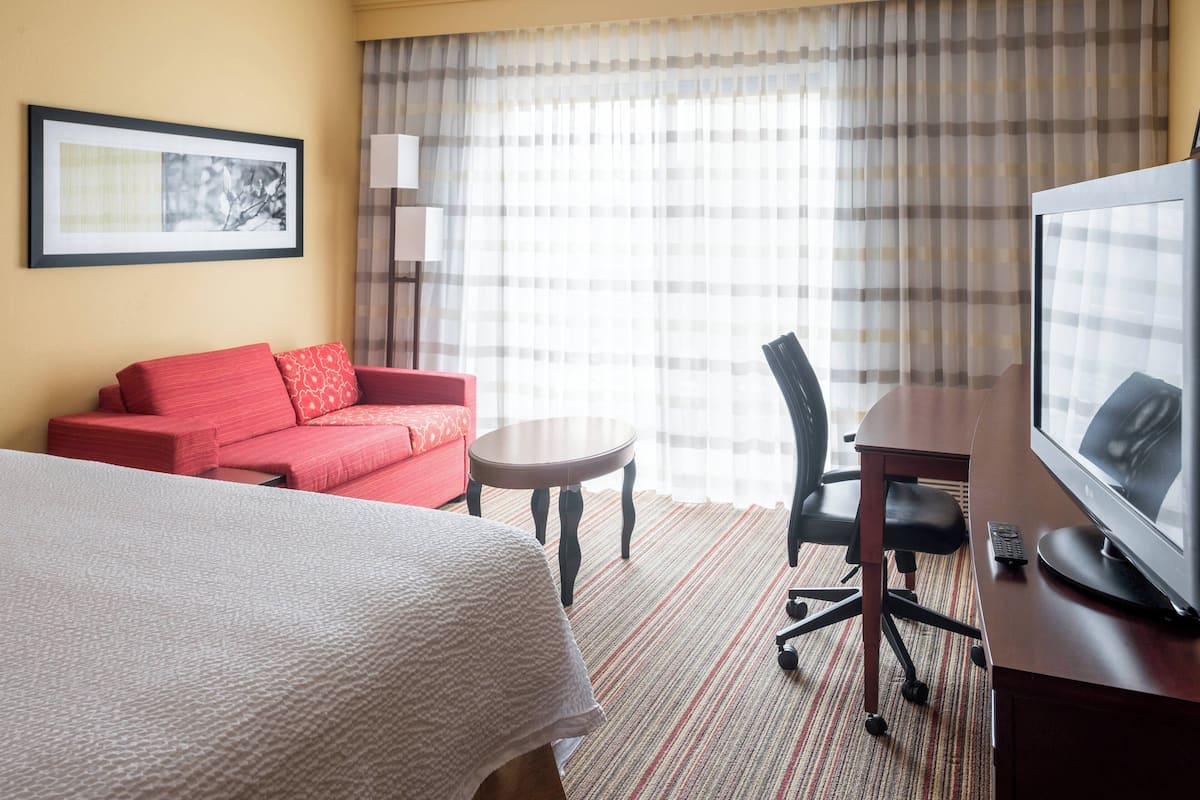 Una camera d'albergo con un letto, un divano rosso, un piccolo tavolino rotondo, una scrivania con un computer e una televisione.