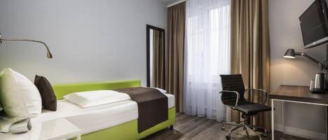 Habitación de hotel con cama, escritorio, silla, televisión y ventana con cortinas.