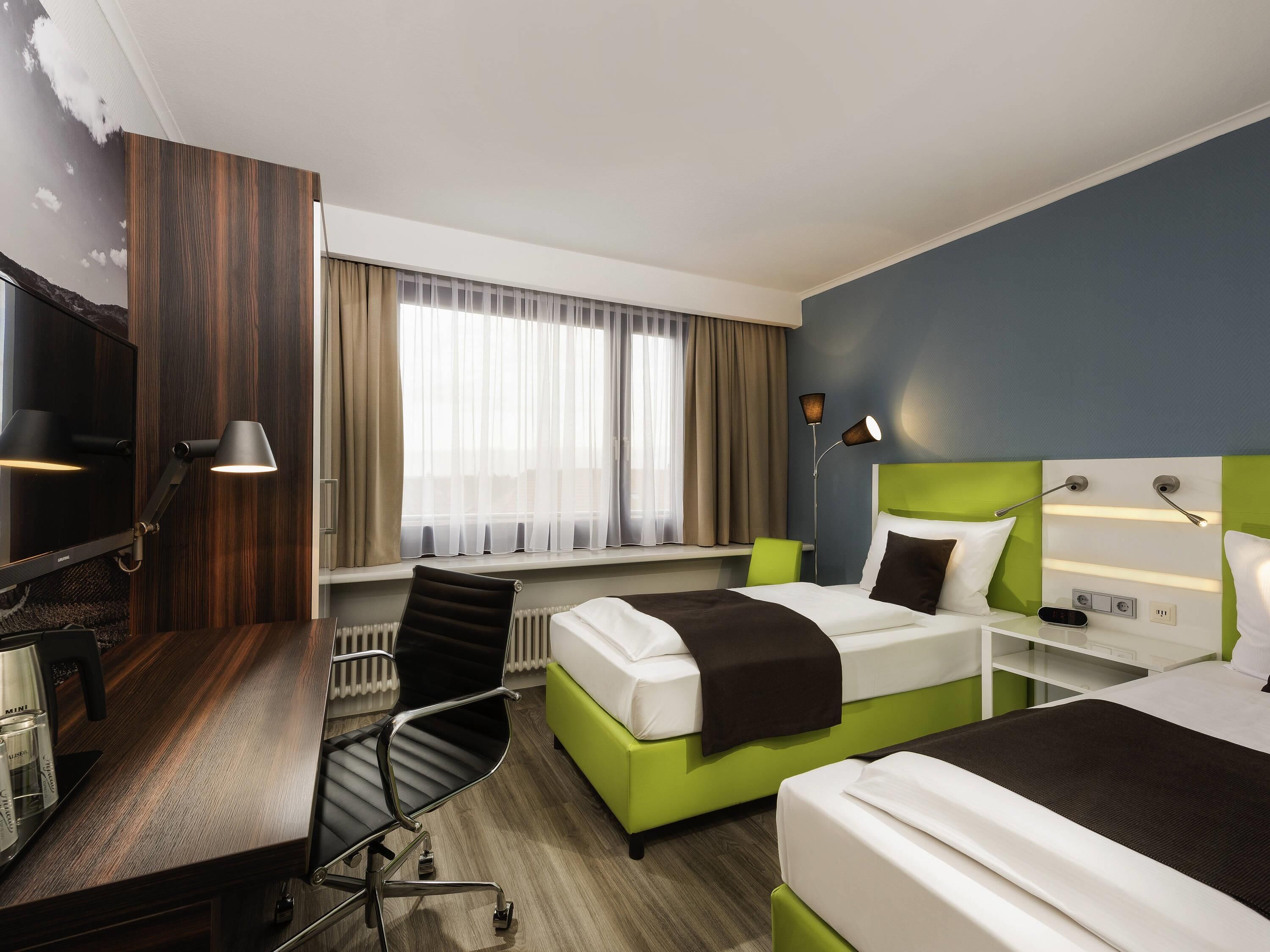 Une chambre d’hôtel avec deux lits, un bureau avec une chaise, une télévision et une fenêtre avec des rideaux.