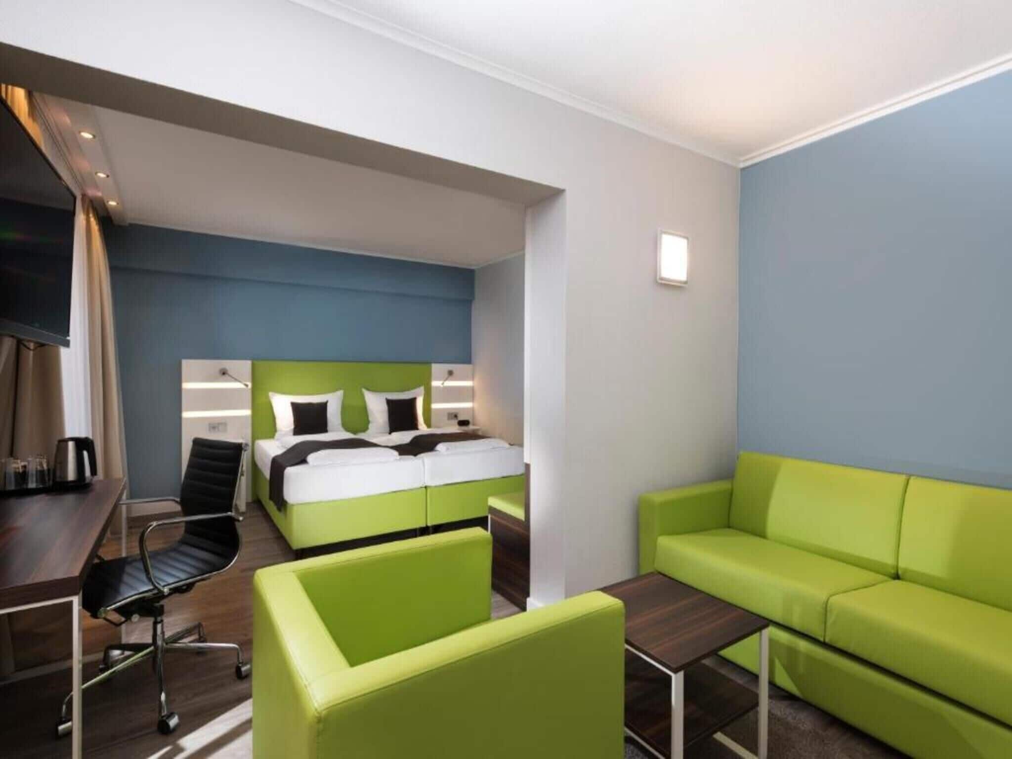 A hotel room with a bed, a desk, a chair, and a green sofa.