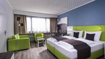 A modern hotel room with a large bed, green armchairs, a desk, and a view of the mountains through the window.