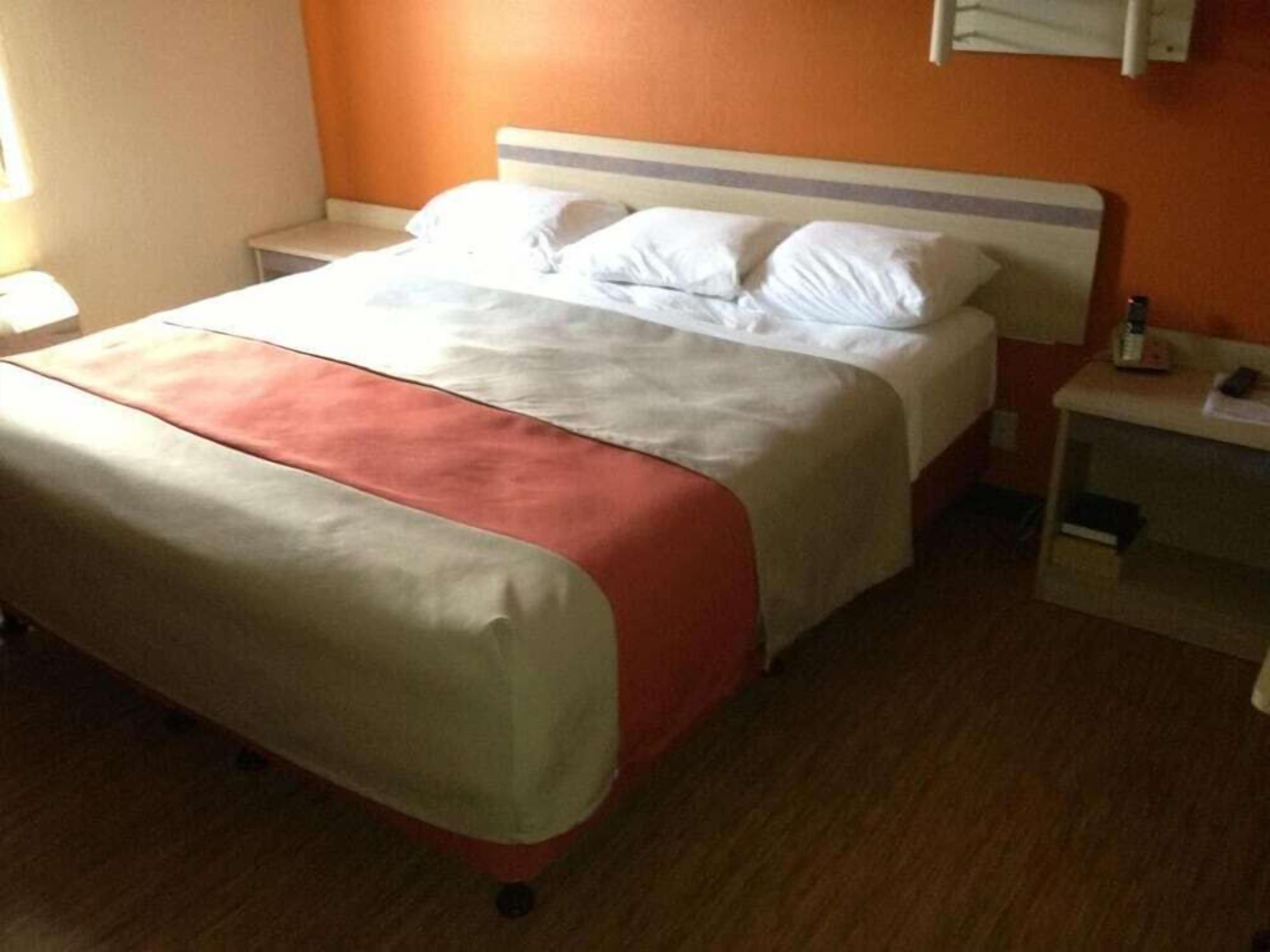 Una cama con una colcha blanca y naranja, una mesita de noche con una lámpara, y una estantería de pared con una botella y un libro.