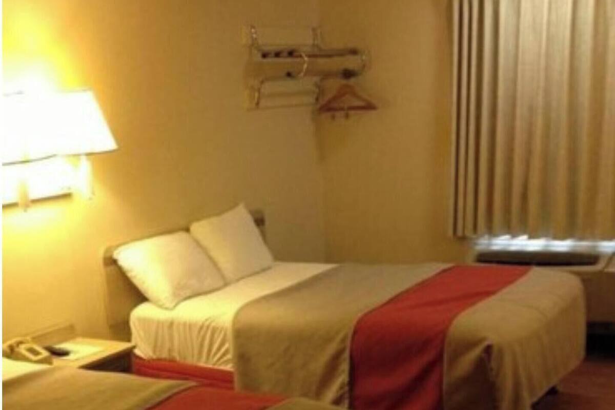 Een hotelkamer met twee bedden, een nachtkastje, een lamp, een kapstok en een raam met gordijnen.