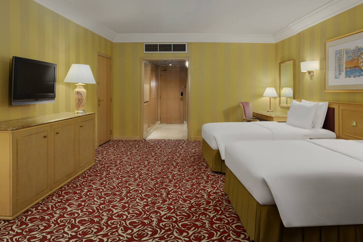 A hotel room with a red and white patterned carpet, a bed, a TV, a desk with a lamp, and a door leading to another room.