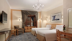 A hotel room with two beds, a desk, a chair, a TV, a chandelier, and a window with curtains.