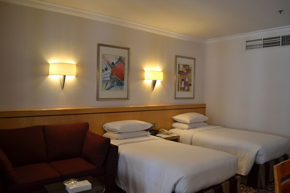 Habitación de hotel con dos camas individuales, un sofá, una mesita y dos lámparas de pared.